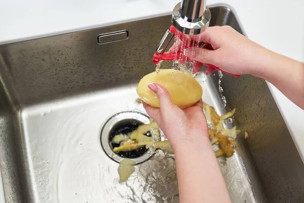 Καλλιεργημένη άποψη των γυναικείων χεριών ξεφλούδισμα πατάτας πάνω από τη μηχανή διάθεσης απορριμμάτων τροφίμων στο νεροχύτη στη σύγχρονη κουζίνα — Φωτογραφία Αρχείου
