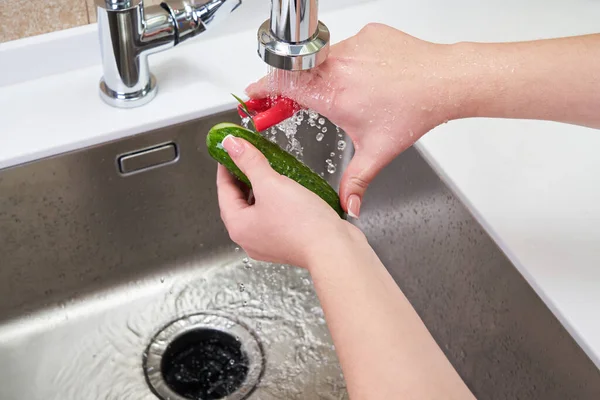 Обрезанный вид женских рук, очищающих огурец над машиной по утилизации пищевых отходов в раковине на современной кухне — стоковое фото