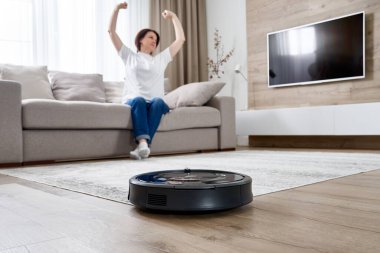 Kadın kanepede dinlenirken robot süpürgesi odayı temizliyor.