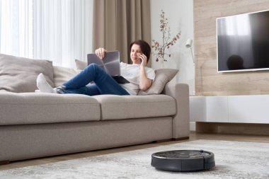 Kadın kanepede dinlenirken robot süpürgesi odayı temizliyor.