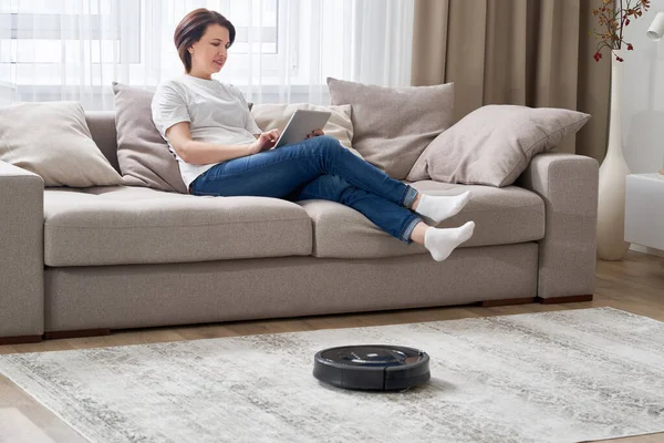 Роботизированный пылесос убирает комнату, пока женщина отдыхает на диване — стоковое фото