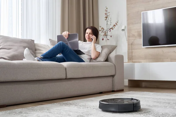 Robotic vacuum cleaner cleaning the room while woman resting on sofa — Stock Photo, Image