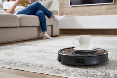 Robotik elektrikli süpürge bir kadına koltukta dinlenirken bir fincan kahve getiriyor.