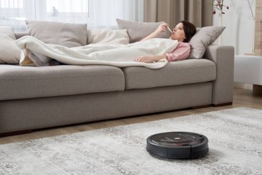 Robot elektrikli süpürge oturma odasındaki halıyı temizlerken kanepede yatan grip hastası kadın.