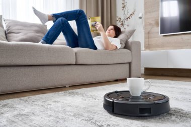 Robotik elektrikli süpürge bir kadına koltukta dinlenirken bir fincan kahve getiriyor.