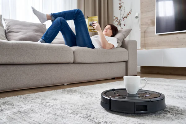 Robotic vacuum cleaner bringing cup of coffee to a woman while she is resting on sofa — Stock Photo, Image