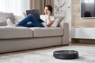 Kadın kanepede dinlenirken robot süpürgesi odayı temizliyor.