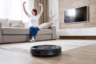 Kadın kanepede dinlenirken robot süpürgesi odayı temizliyor.