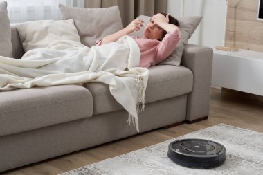 Robot elektrikli süpürge oturma odasındaki halıyı temizlerken kanepede yatan grip hastası kadın.