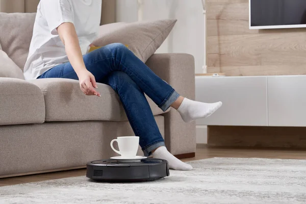 Robotic vacuum cleaner bringing cup of coffee to a woman while she is resting on sofa — Stock Photo, Image