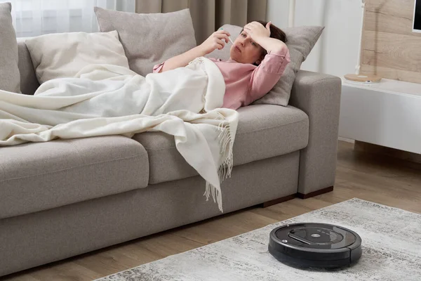 Женщина с гриппом лежит на диване, пока робот пылесос чистит ковер в гостиной — стоковое фото