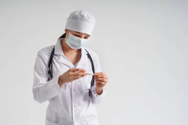 Врач в маске смотрит на измерение на медицинском термометре на белом фоне с копировальным пространством — стоковое фото
