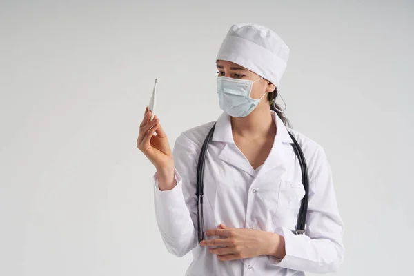 Läkare bär ansiktsmask tittar på mätning på medicinsk termometer över vit bakgrund — Stockfoto