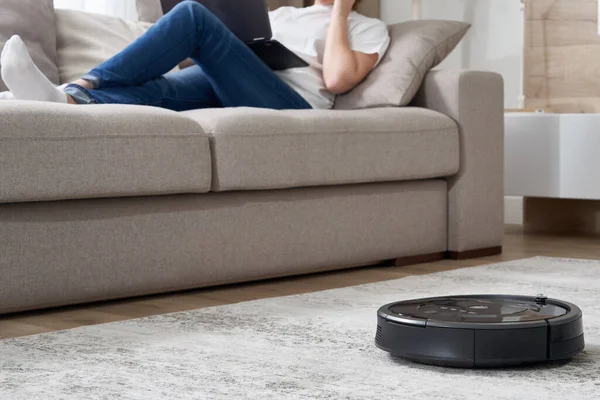 Kadın kanepede dinlenirken robot süpürgesi odayı temizliyor.