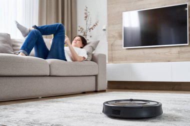 Kadın kanepede dinlenirken robot süpürgesi odayı temizliyor.