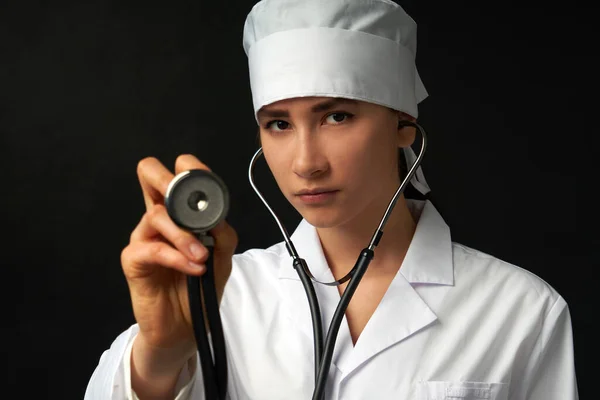 Cropped view of young female doctor holding stethoscope hanging on her neck, studio shot over black background with copy space — Stock Photo, Image