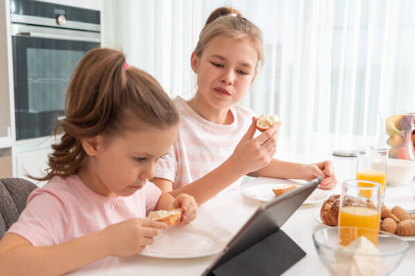 Two sisters having breakfast and watching cartoons on tablet together, happy family concept