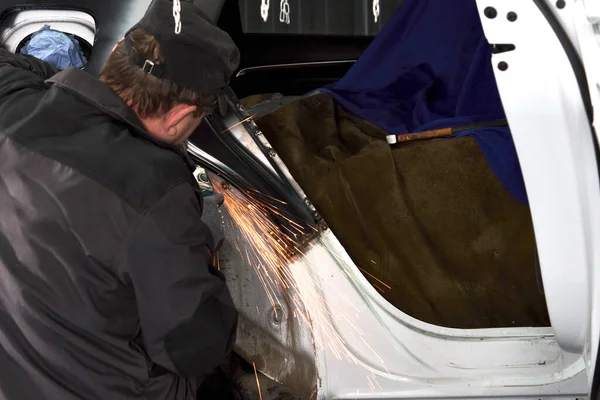 Petugas perbaikan memperbaiki mobil yang rusak. Bekerja dengan penggiling sudut untuk memperbaiki tubuh logam. — Stok Foto
