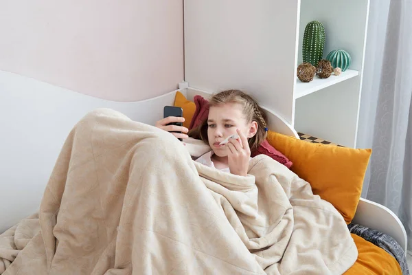 Hasta genç kız cep telefonuyla termometreyle yatakta yatıyor. — Stok fotoğraf