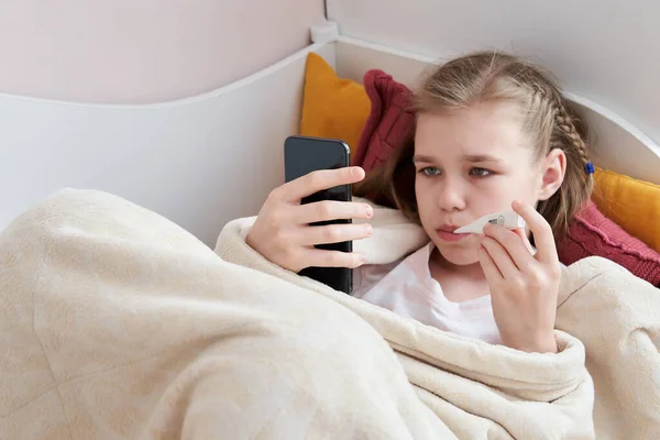 Hasta genç kız cep telefonuyla termometreyle yatakta yatıyor. — Stok fotoğraf