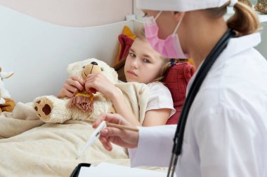 Doktor termometreyi tutuyor ve hasta küçük kızı kontrol ediyor. Ateşli küçük kız yatakta yatıyor.