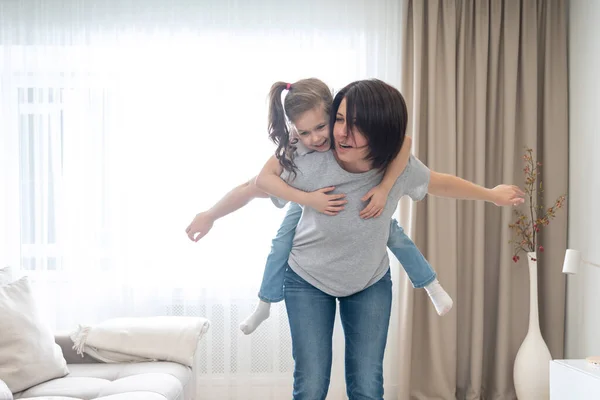 Gelukkig moeder geven vrolijke piggyback rit naar haar dochter, plezier hebben thuis, alleenstaande moeder gelukkig familie concept — Stockfoto