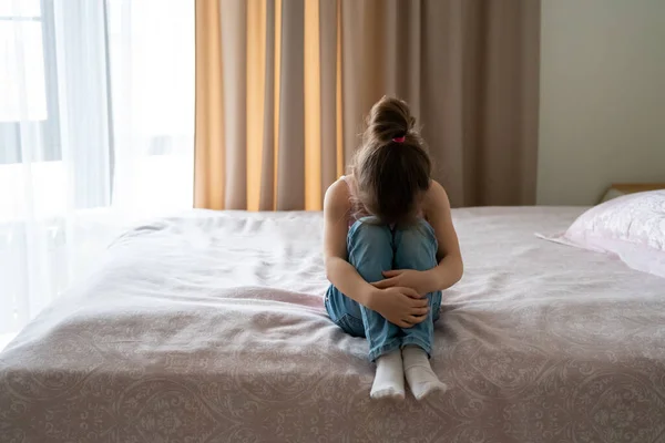 Une petite fille triste assise sur un lit, vue de derrière — Photo