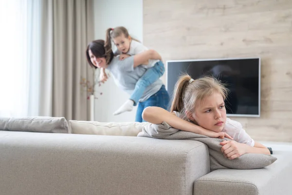 Preteenage sad girl sitting on a couch while mother having fun with sister, jealousy concept — Stock Photo, Image