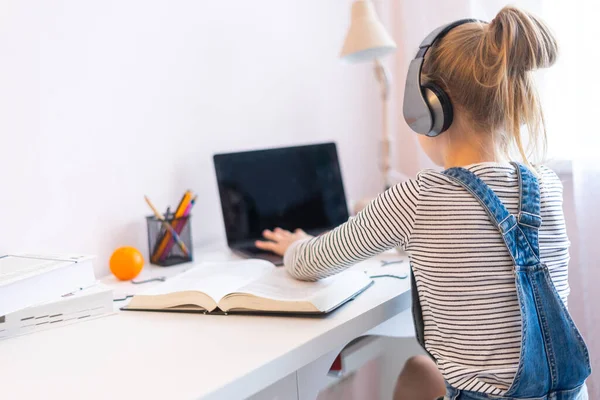 Портрет дівчини-підлітка, яка навчається онлайн з навушниками та ноутбуком, роблячи нотатки в блокноті, сидячи за своїм столом вдома, роблячи домашнє завдання — стокове фото