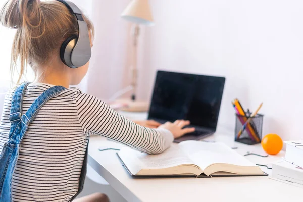 Портрет дівчини-підлітка, яка навчається онлайн з навушниками та ноутбуком, роблячи нотатки в блокноті, сидячи за своїм столом вдома, роблячи домашнє завдання — стокове фото