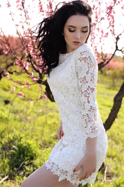 Великолепная молодая женщина в элегантном платье позирует в саду с цветущими персиковыми деревьями — стоковое фото