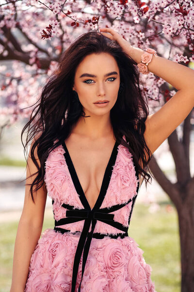 fashion outdoor photo of beautiful woman with dark hair in elegant dress with accessories posing among blossoming peach trees