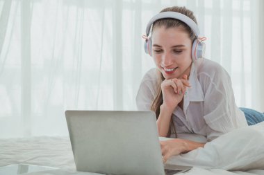 Kadın dizüstü bilgisayarı kullanarak yatakta dinleniyor ve müzik dinliyor.