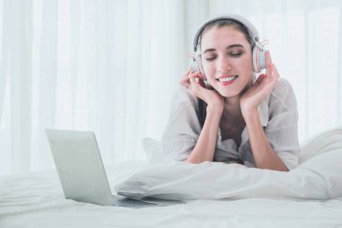 Güzel bir kadın yatakta dizüstü bilgisayar kullanırken dinleniyor ve müzik dinliyor.