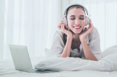 Genç bir kadın yatakta dizüstü bilgisayar kullanırken dinleniyor ve müzik dinliyor.