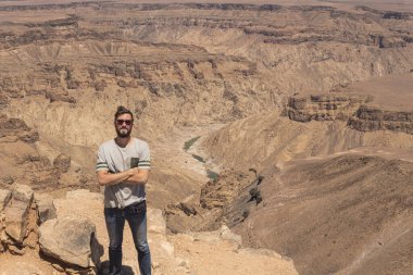 Genç adam Namibya 'daki Fish River Kanyonu' nda poz veriyor. Kolları bağlı sakallı bir adam. Çölde yol