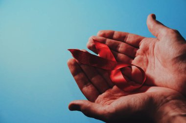 Aralık ayındaki Dünya Yardım Günü kampanyası. Kırmızı kurdele farkındalığını mavi arka plandaki adamın ellerine yakın tut. Hiv 'de yaşayanlara farkındalığın ve desteğin sembolü. Boşluğu kopyala.