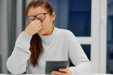 Elektronik kitap okurken burun masajı yapan gözlüklü ve örgü ören beyaz bir kadın. Baş ağrısı, stres, tansiyon sorunu, migren.