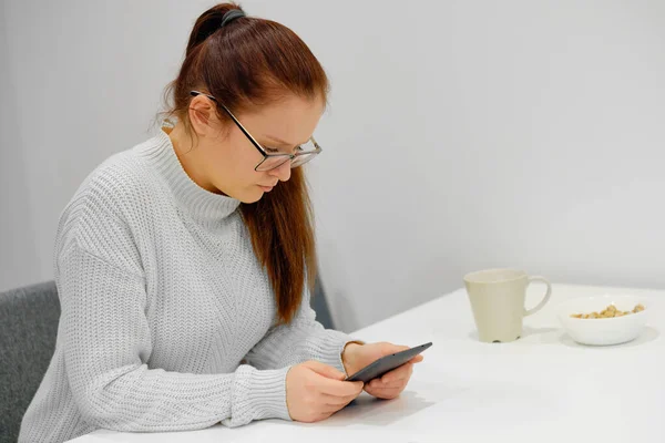 Young caucasian woman in gray knitted sweater seats at the table in modern light interior and reading an electronic book. Concept of the education, exam preparation, reading