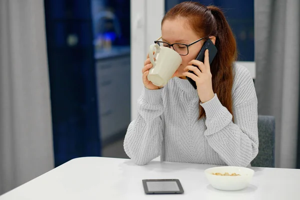Gri örgü süveterli genç beyaz kadın modern aydınlık iç mekânda masada oturuyor ve cep telefonuyla konuşurken kahve içiyor.
