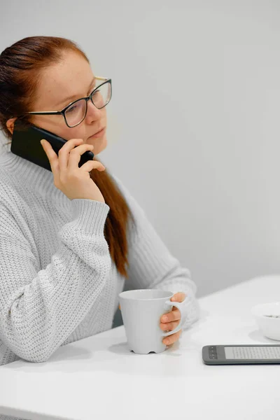 Gri örgü süveterli genç beyaz kadın modern aydınlık iç mekânda masada oturuyor ve cep telefonuyla konuşurken kahve içiyor.
