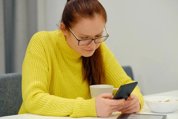 Young caucasian woman in yellow knitted sweater smilimng while seats at the table in modern light interior and checking the mobile phone messages