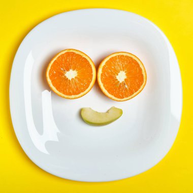 Beyaz tabaktaki iki portakal ve bir elma diliminin üst görüntüsü gülümserken, parlak sarı arka planda yatıyor. Sağlıklı yemek, detoks, diyet konsepti