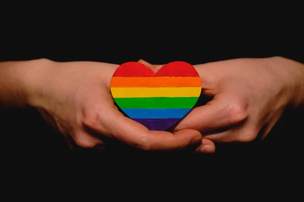Brazos femeninos que sostienen el corazón coloreado en colores de orgullo LGBT sobre el fondo oscuro. Concepto del Día Internacional contra la Homofobia concepto, igualdad de género, feminismo, seguridad social —  Fotos de Stock