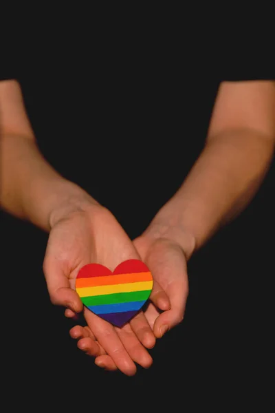 Brazos femeninos que sostienen el corazón coloreado en colores de orgullo LGBT sobre el fondo oscuro. Concepto del Día Internacional contra la Homofobia concepto, igualdad de género, feminismo, seguridad social —  Fotos de Stock