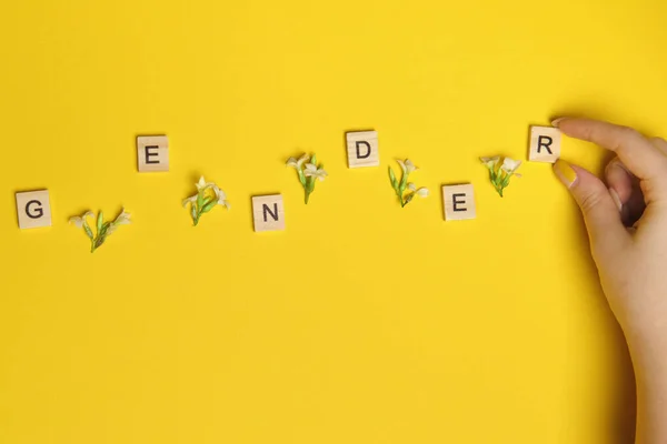 Word gender depicted by using wooden blocks and flowers on yellow bee background with woman hand pointing on it