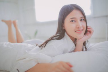 Beyaz tişörtlü Asyalı kadının beyaz çarşafların üzerinde sabah ışığında çenesini tutuşu. Mutlu yaşam, rahatlama, rahatlama kavramı.