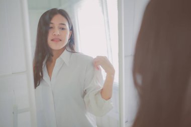 Beyaz gömlekli Asyalı kadın aynanın yanında poz veriyor..