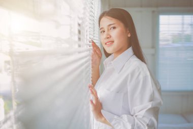 Büyük beden beyaz gömlekli Asyalı bir kadın güneşli bir günde pencerenin yanında güneşli panjurlarla poz veriyor.. 