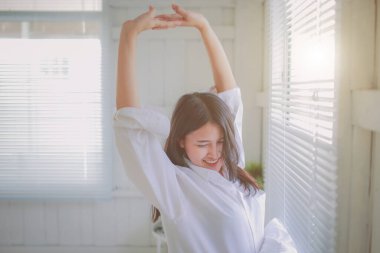 Mutlu Asyalı kadın portresi, sabah yatak odasında uyandıktan sonra, perdelerin ardından güneş ışığıyla geriniyor..  